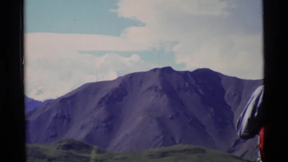Alaska mountain landscape in a 1977 home-movie frame, with a cropped hiker visible at the right edge.