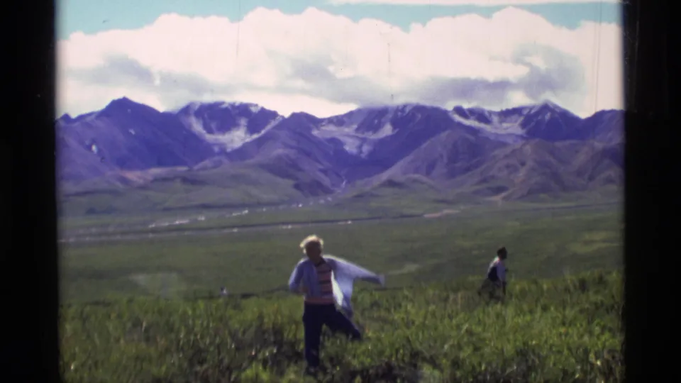 1977 Alaska home-movie frame showing two hikers moving through open grass below distant mountains and clouds.