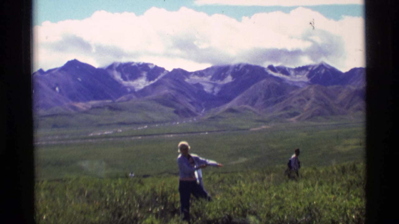 1977 Alaska 8mm frame showing a person in a light jacket crossing grassy tundra below snow-streaked mountains.