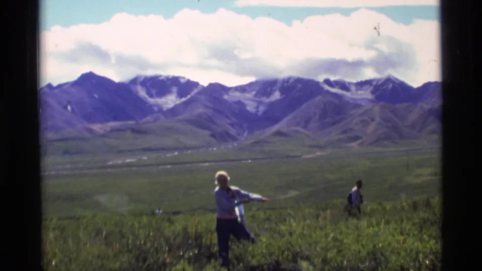 1977 Alaska 8mm frame showing a person in a light jacket crossing grassy tundra below snow-streaked mountains.