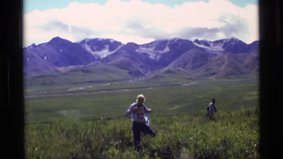 Home-movie frame from Alaska showing a person in striped clothing on a grassy slope with another figure and mountains behind.