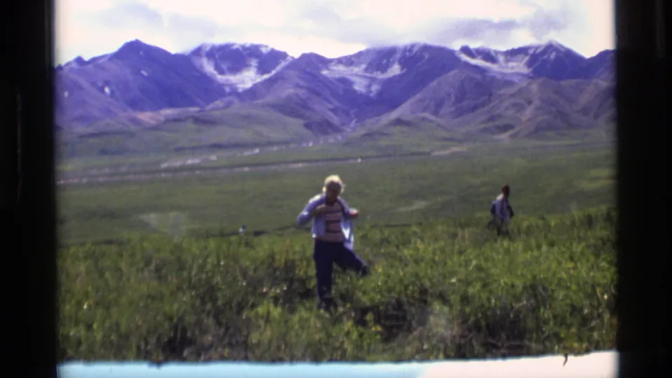 1977 Alaska 8mm still with a person in a striped shirt and light jacket in grass, backed by a wide valley and mountains.