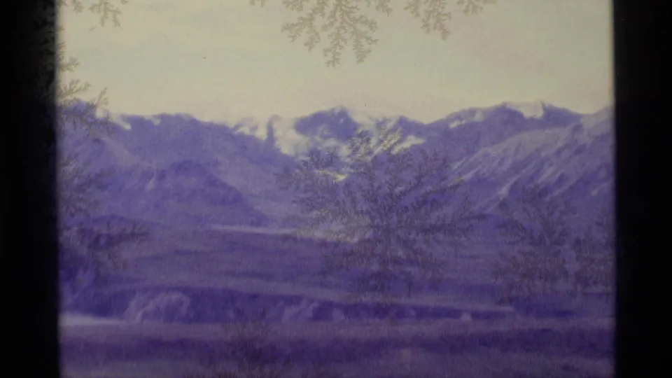 1977 Alaska 8mm frame of snow-streaked mountain ridges viewed through frost-patterned glass and a dark vertical window divider.