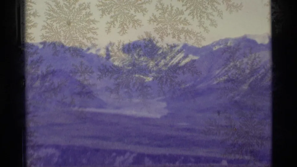 1977 Alaska 8mm frame with dense frost on glass, dark window borders, snow-covered mountain slopes, and a soft purple-blue cast.