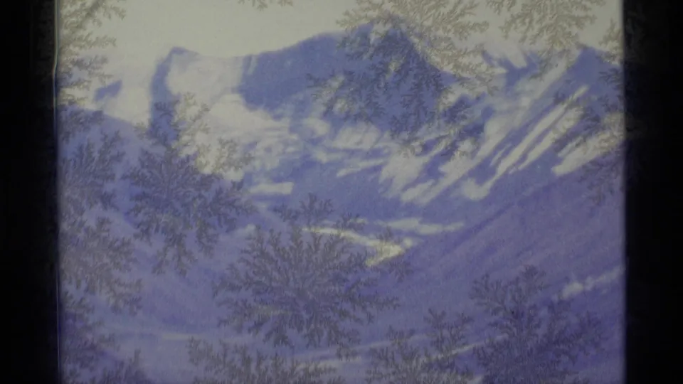 Snowy Alaskan mountain peaks appear behind delicate frost on glass in this 1977 home-movie frame.
