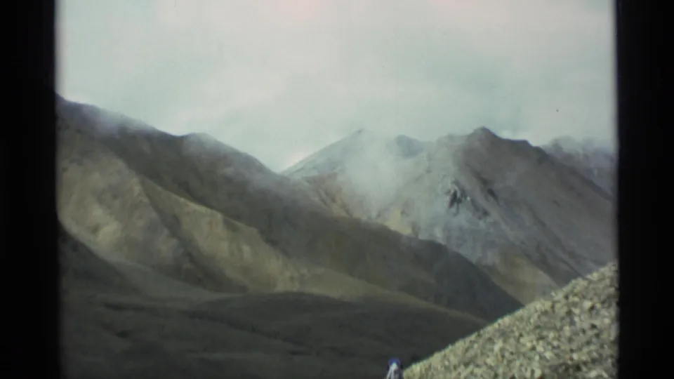 1977 Alaska home-movie frame with a misty mountain range and a small hiker partly visible near the rocky foreground.