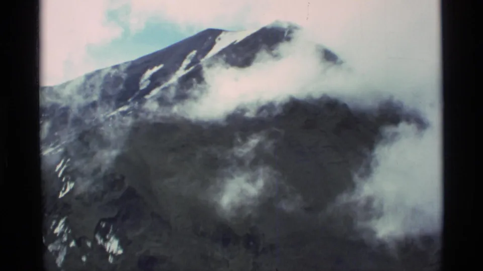 1977 Alaska 8mm frame of a dark, snow-streaked mountain peak partly obscured by drifting cloud.