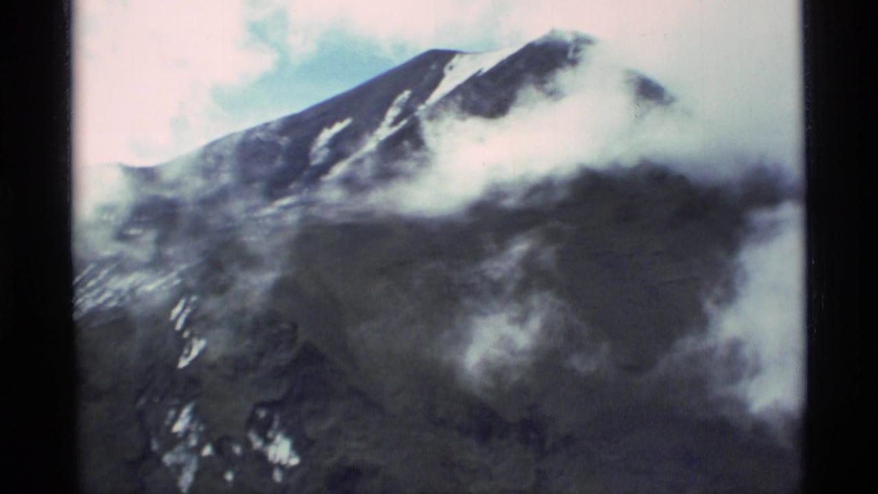 1977 Alaska 8mm frame showing a dark mountain peak with patches of snow, low clouds crossing the slope, pale sky, and black film borders.