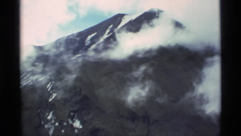 1977 Alaska 8mm frame showing a dark mountain peak with patches of snow, low clouds crossing the slope, pale sky, and black film borders.
