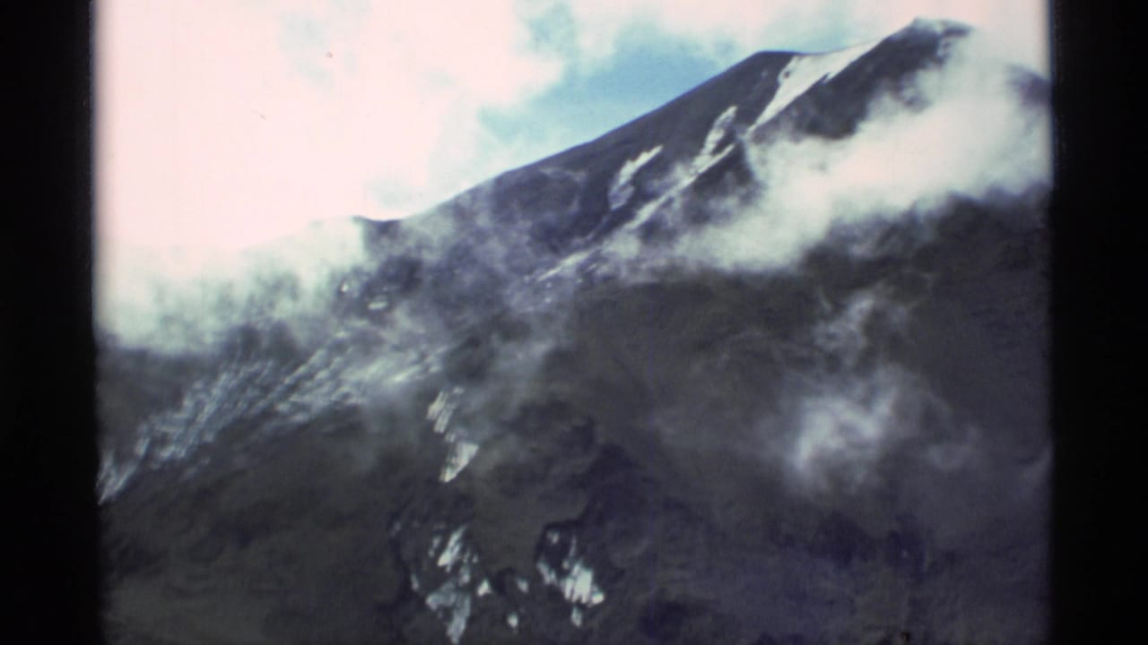 1977 Alaska 8mm frame with a high mountain ridge, scattered snow, gray cloud bands, pale sky, and dark edge borders from the film frame.