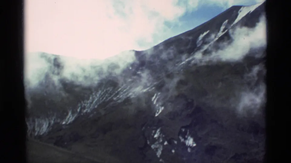 1977 Alaska 8mm frame showing a rugged mountain slope with scattered snow, broad cloud cover, pale sky, soft focus, and visible film-frame borders.