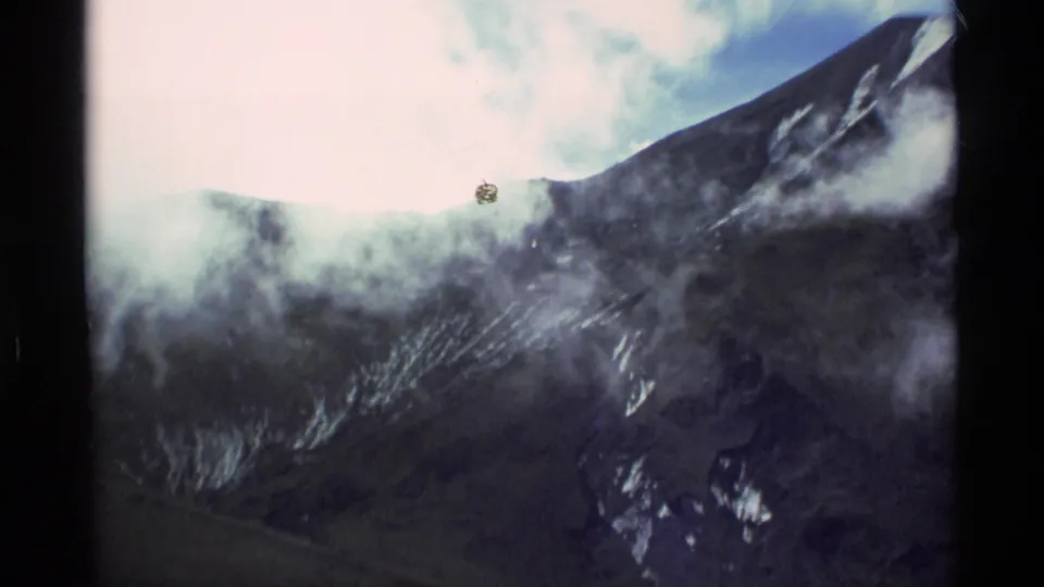 1977 Alaska 8mm frame showing cloud and mist over a snow-marked mountain slope, with pale sky, dark film borders, and a small visible dust or scan speck near the upper center.