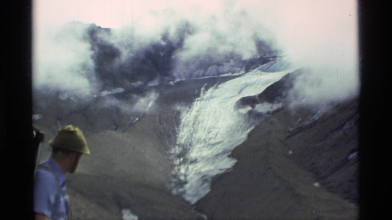 1977 Alaska 8mm frame showing a man at left observing steam or smoke above an ice-covered mountain slope.