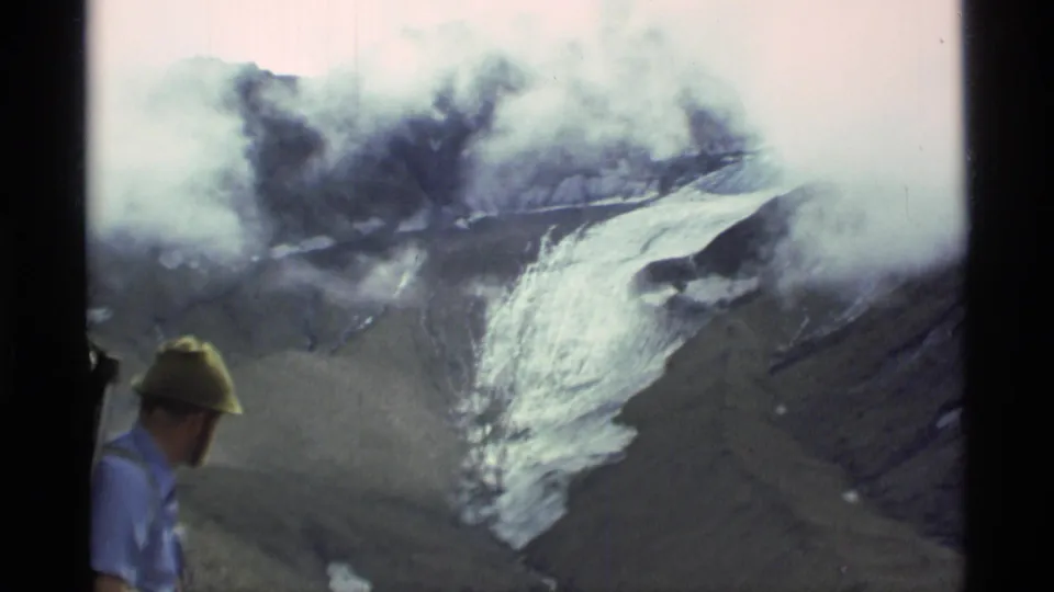 1977 Alaska 8mm frame showing a man at left observing steam or smoke above an ice-covered mountain slope.