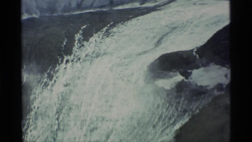 Closer 8mm view of bright ice and snow crossing a dark Alaska mountain slope in the 1977 travel footage.