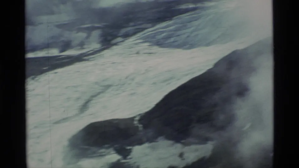 Soft, slightly blurred 1977 Alaska frame showing ice spread across a dark mountain slope with pale vapor nearby.