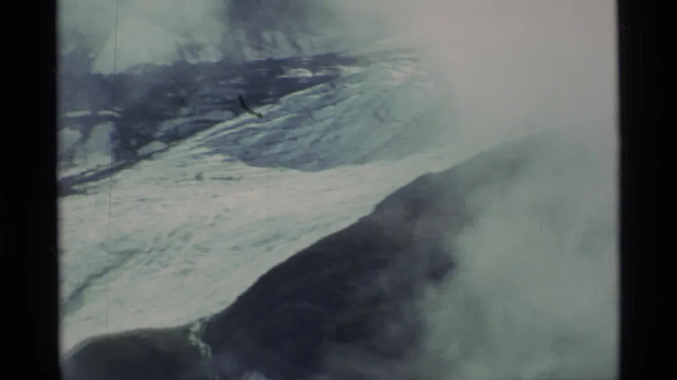 1977 Alaska 8mm aerial frame showing pale ice fields, dark ridges, and mist over rugged mountain terrain.