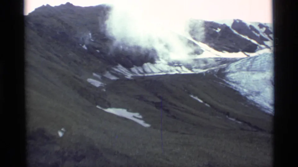Aerial 8mm view in Alaska showing a dark mountain ridge, snow patches, and cloud or mist moving across the peaks.