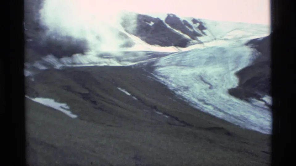 1977 Alaska aerial home-movie frame with an icy slope, dark rocky ridge, low mist, and green-gray mountain terrain.
