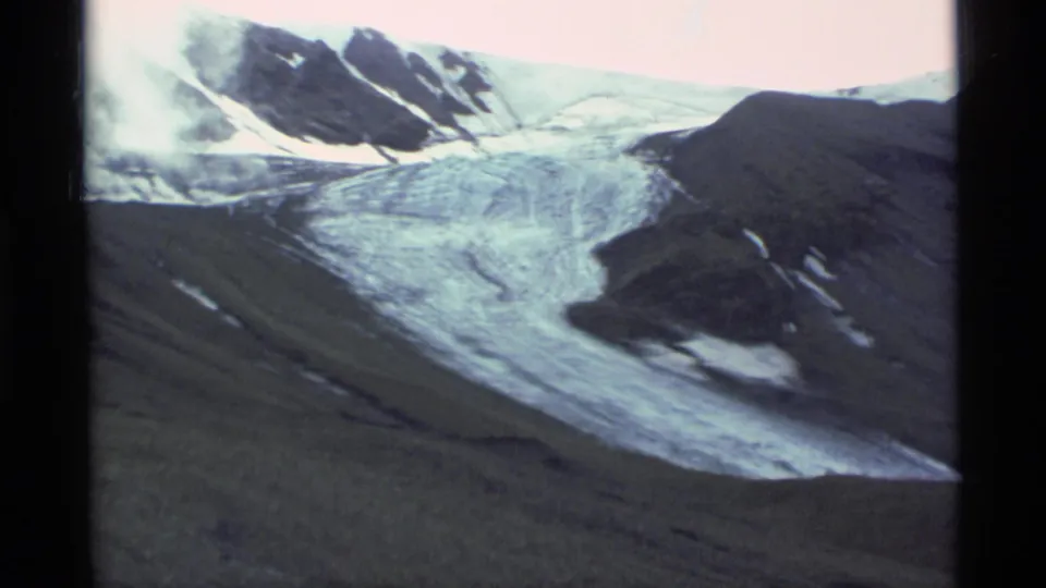 Vintage 8mm aerial frame from Alaska showing an icy mountain valley, dark ridges, snow patches, and pale sky.