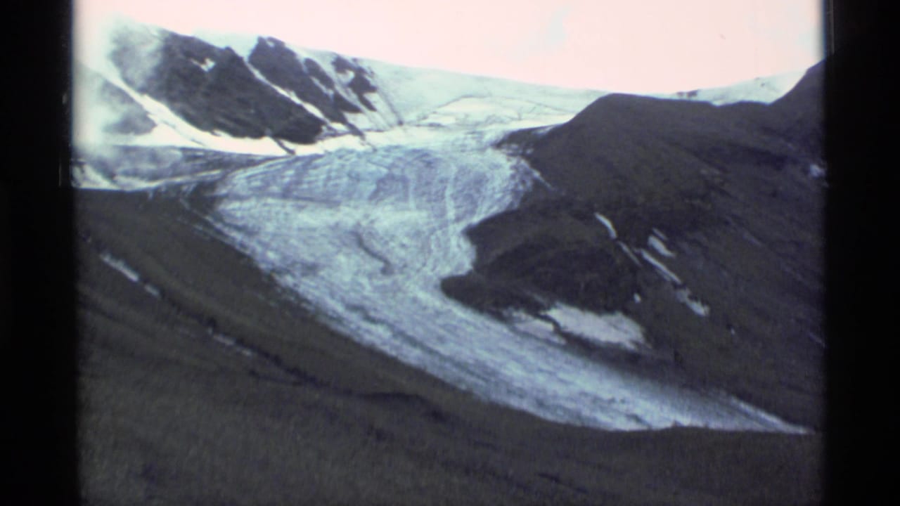 1977 Alaska home-movie view of a glacier curving through rocky mountain terrain with snow patches and visible film borders.
