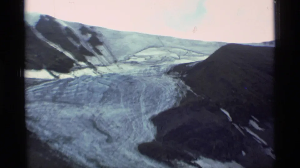Alaska mountain landscape with a broad glacier, dark ridges, snow traces, and faded 8mm color from a handheld viewpoint.
