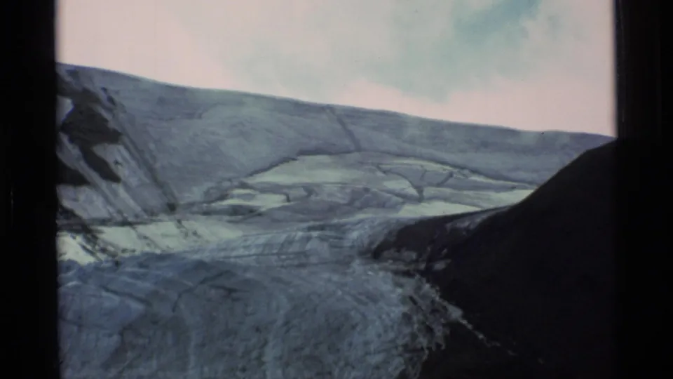 Faded 8mm Alaska landscape showing a glacier receding between dark valley walls beneath a pale, lightly clouded sky.
