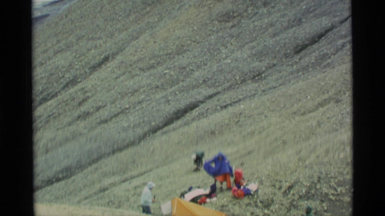 Hikers and gear on a rocky mountain slope with a bright tent nearby.