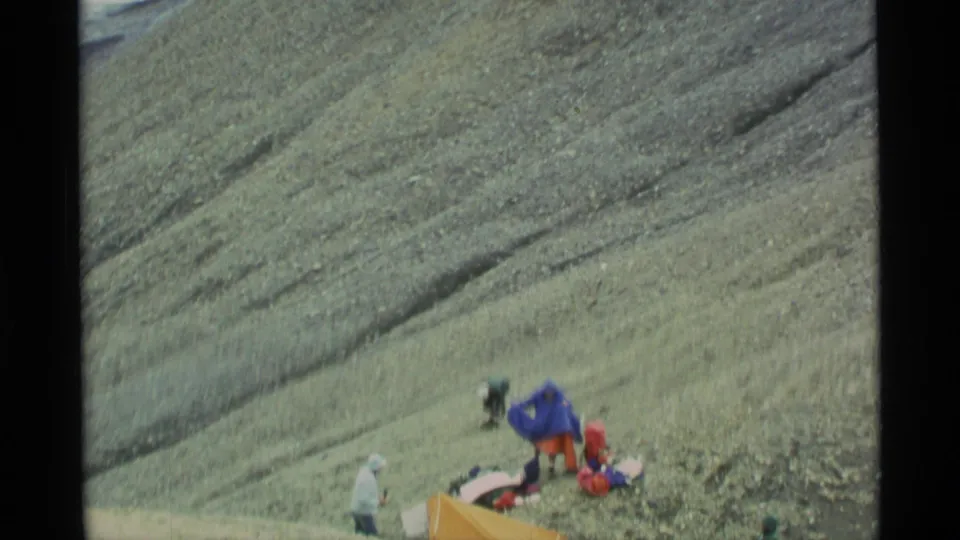 1977 Alaska 8mm frame showing hikers paused beside gear and a bright tent on a steep rocky slope.