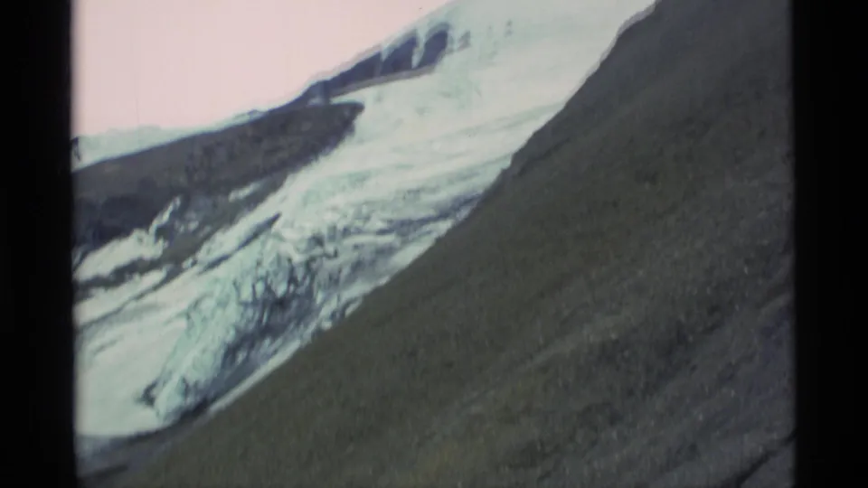 1977 Alaska 8mm frame showing glacier ice, snowfields, and a dark rocky slope in the mountains.