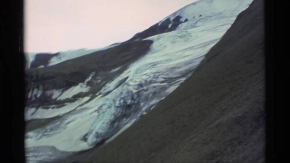 1977 Alaska 8mm frame of a glacier sweeping between dark mountain ridges and a steep rocky slope.
