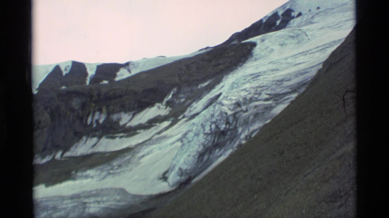 1977 Alaska home-movie frame showing glacier ice, dark rocky slopes, and snow patches under a pale sky.
