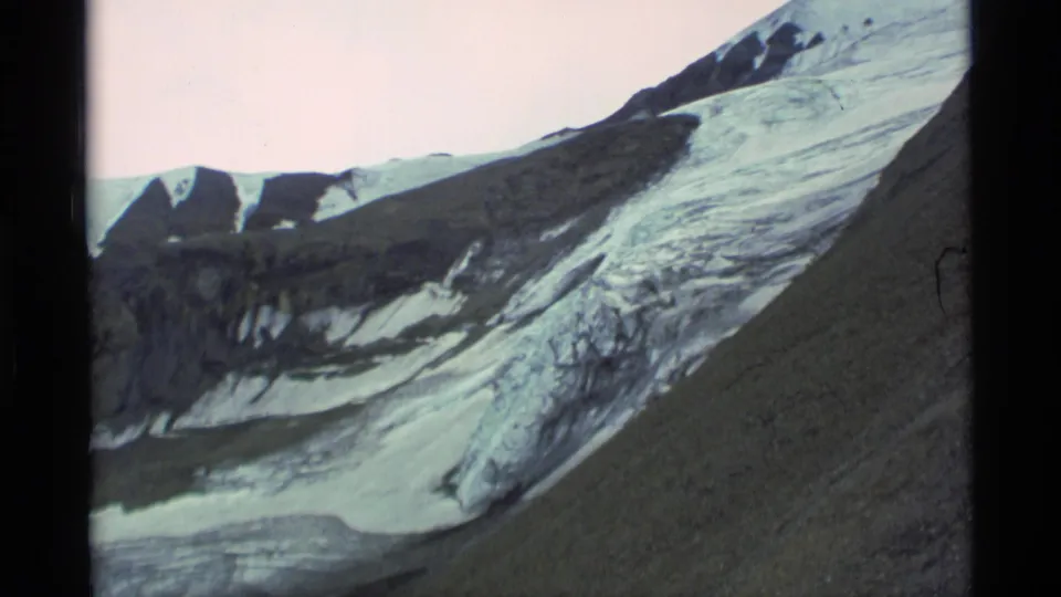 1977 Alaska home-movie frame showing glacier ice, dark rocky slopes, and snow patches under a pale sky.