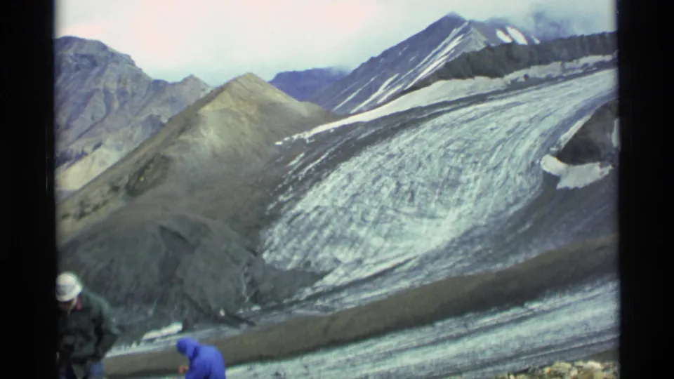 1977 Alaska home-movie frame with two distant people near gear in front of glacier ice and steep mountain slopes.