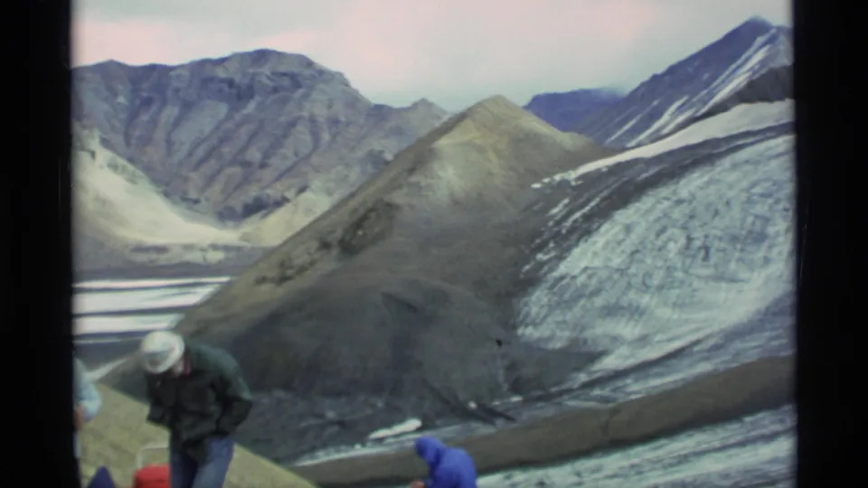 Alaska 1977 frame showing distant people in winter clothing beside a red container, with glacier ice and ridges behind them.