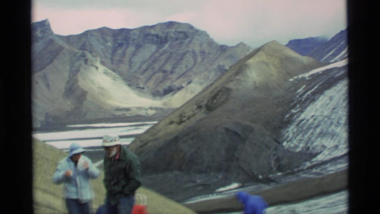 1977 Alaska home-movie frame showing two hikers in hats and jackets on a rocky alpine slope above a broad mountain valley.