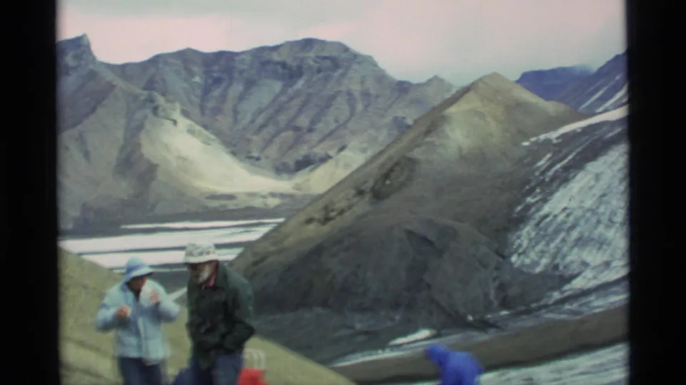 1977 Alaska home-movie frame showing two hikers in hats and jackets on a rocky alpine slope above a broad mountain valley.