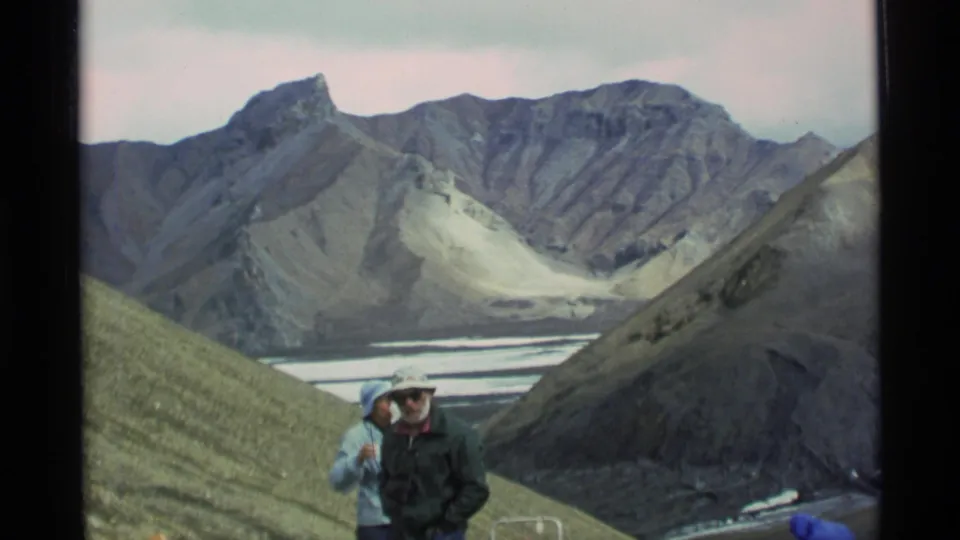 1977 Alaska 8mm frame with hikers in cold-weather clothing standing before steep ridges, snow patches, and a distant valley.