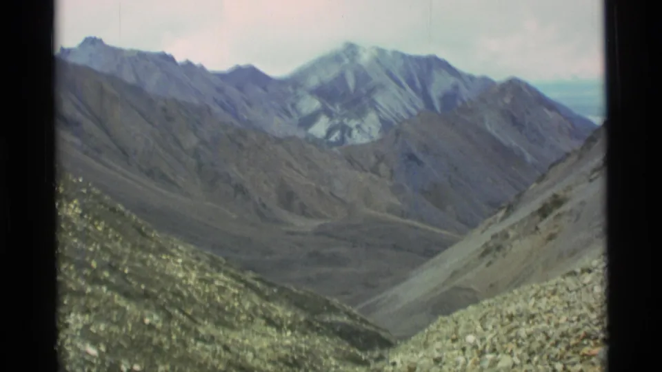 1977 Alaska home-movie landscape frame showing rugged mountain ridges, a rocky valley, and pale snow or ice on distant peaks.