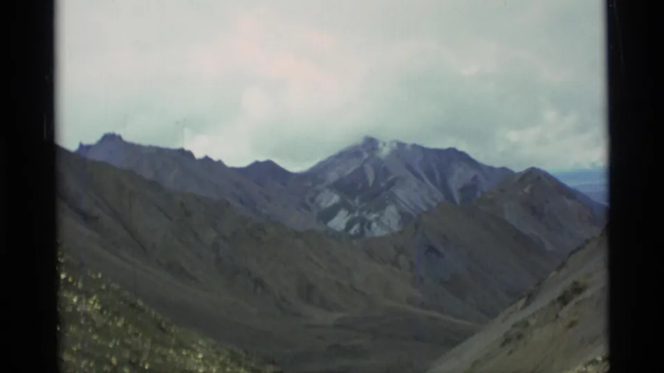 1977 Alaska 8mm landscape frame of rugged ridges and valley terrain under cloud cover, viewed from a high rocky slope.