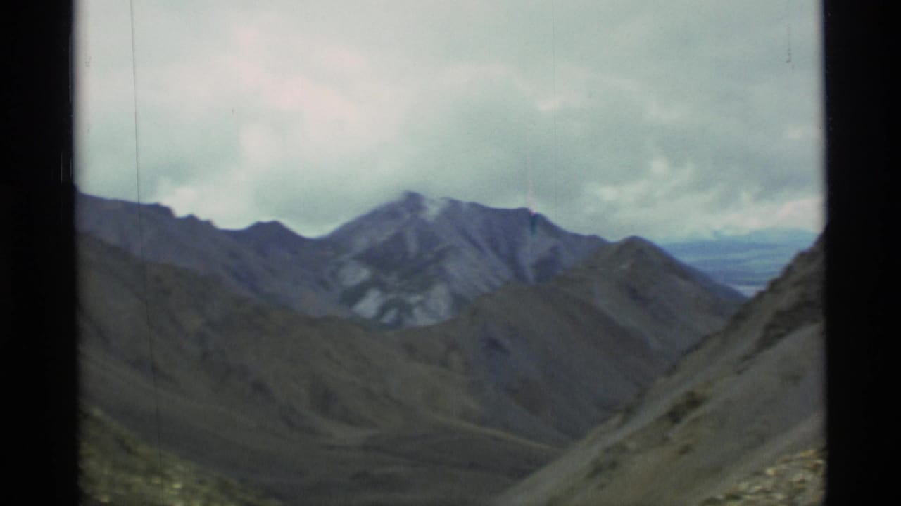 1977 Alaska 8mm frame showing rugged mountain ridges, barren slopes, and a distant valley beneath overcast clouds.