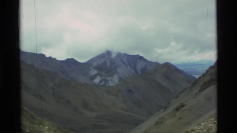 1977 Alaska 8mm frame showing rugged mountain ridges, barren slopes, and a distant valley beneath overcast clouds.