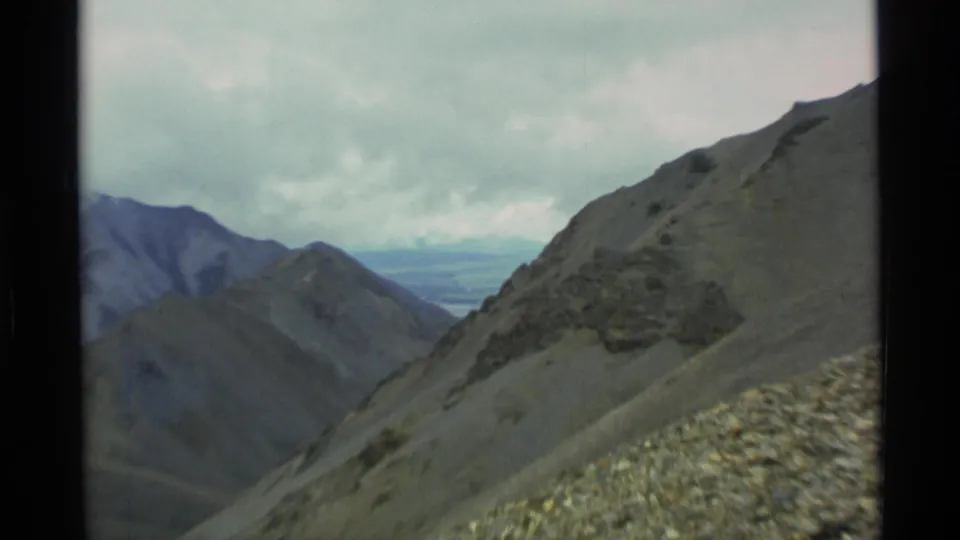 1977 Alaska home-movie view across steep alpine slopes toward layered mountains and a pale distant valley.