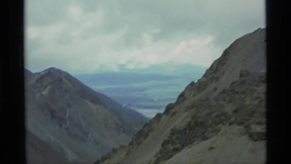 1977 Alaska 8mm frame looking past a rocky mountainside toward a hazy valley under low clouds.