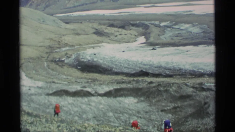 1977 Alaska home-movie frame with distant hikers in red and blue jackets crossing rocky alpine terrain near snow or ice.