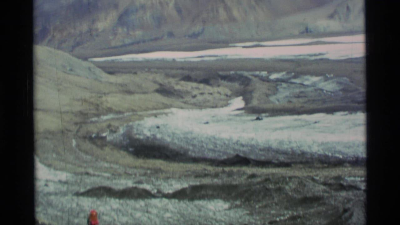 1977 Alaska 8mm frame showing a rugged mountain valley, pale snow or ice, and a distant hiker in a red jacket for scale.
