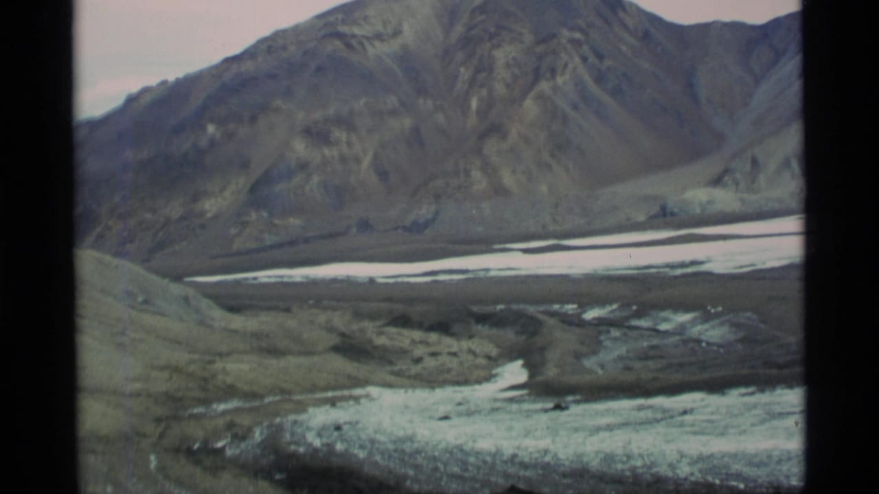 1977 Alaska home-movie view of steep mountain terrain and pale snow or ice across a remote valley floor.