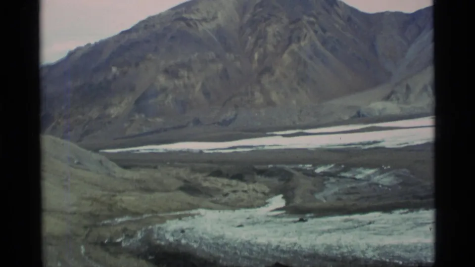 1977 Alaska home-movie view of steep mountain terrain and pale snow or ice across a remote valley floor.