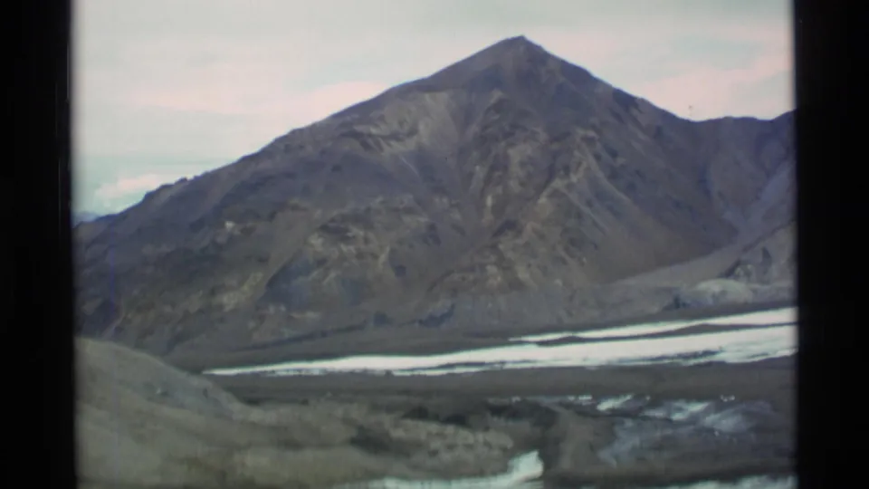 1977 Alaska 8mm frame with a dark mountain peak, barren slopes, and snow or ice crossing the lower valley.