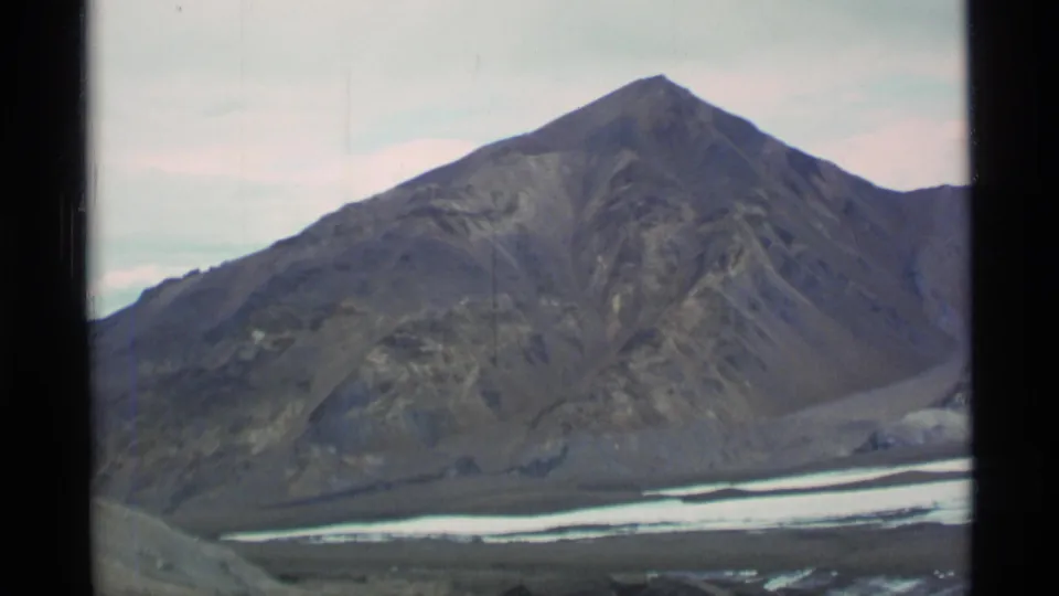 1977 Alaska home-movie frame showing a steep mountain peak over a remote valley with pale snow or ice below.